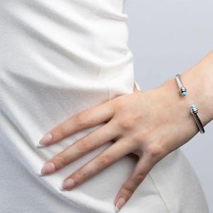 A person with a French manicure wearing a silver twisted cable cuff bracelet with small turquoise beads on their wrist.