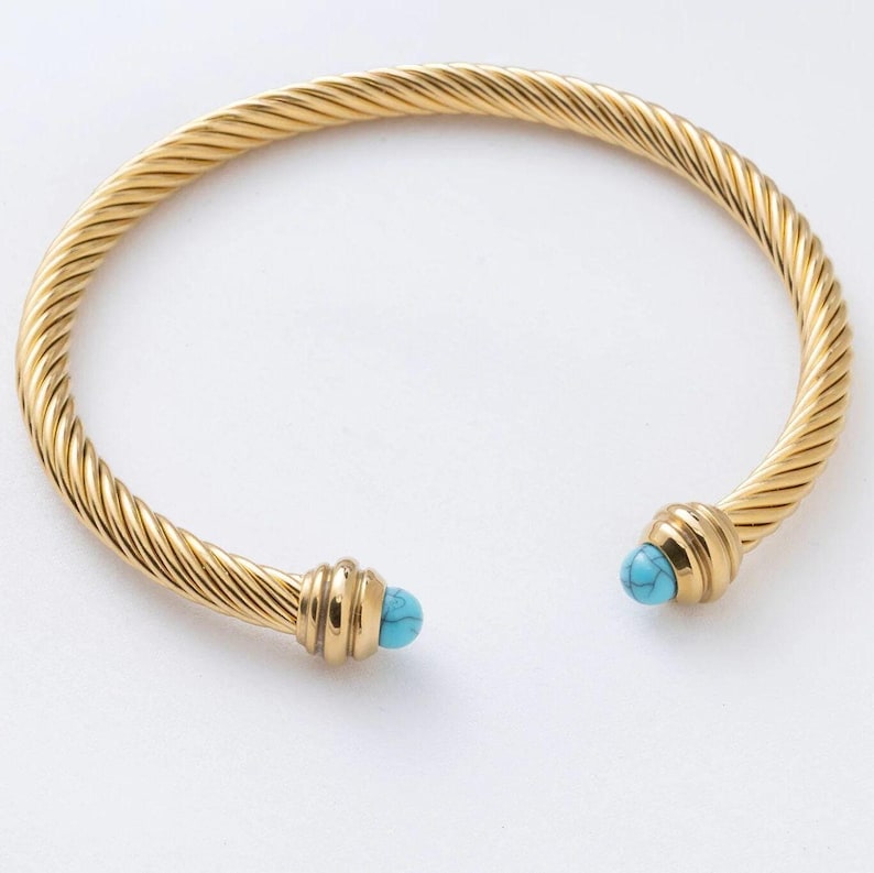 A close-up of a gold twisted cable cuff bracelet with bright turquoise cabochon ends on a white surface.