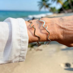 May include: Two silver-toned bracelets featuring a continuous wavy, serpentine design. The metal surface has a subtle hammered texture, creating a reflective finish. The bracelets are open-ended, showcasing a fluid, rhythmic shape that wraps around the wrist.