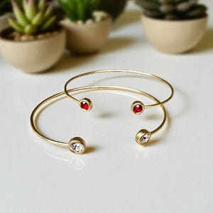 May include: Two gold-colored open cuff bracelets. One bracelet has two red gemstones, and the other has one clear gemstone and one red gemstone. The bracelets are displayed on a white surface with small potted plants in the background.
