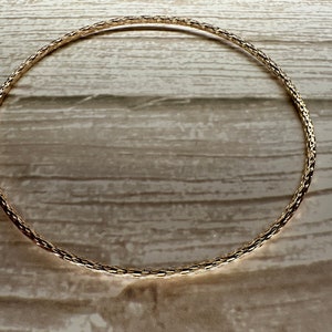 May include: A gold-toned chain bracelet with a delicate, woven design. The bracelet is lying flat on a wooden surface.