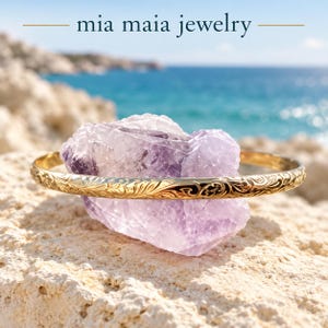 May include: A gold bangle bracelet with an intricate engraved floral pattern resting on a large, translucent purple amethyst crystal. The text 'mia maia jewelry' is displayed in dark blue lettering at the top of the image.