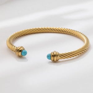 A single gold twisted cable cuff bracelet with turquoise ball ends resting on a white background.