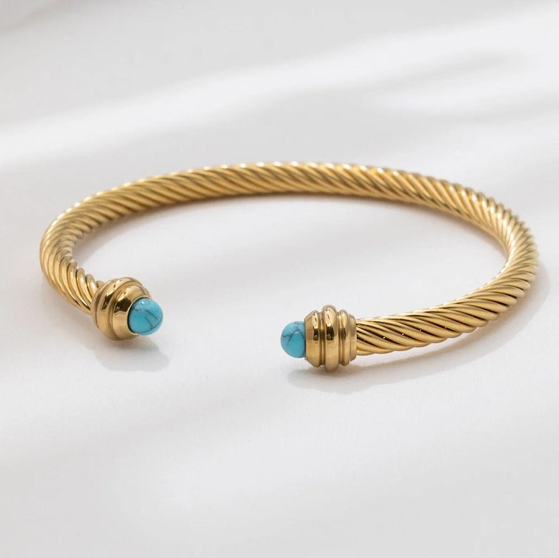 A single gold twisted cable cuff bracelet with turquoise ball ends resting on a white background.