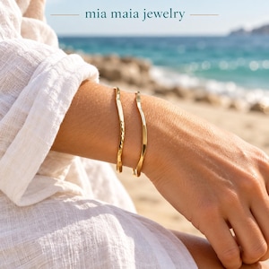 May include: Two gold-toned metal bracelets with a wavy, irregular design. One bracelet features a hammered, textured surface, while the other has a smooth, polished finish. The text "mia maia jewelry" is visible at the top of the image.