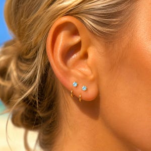 May include: A pair of gold hoop earrings and two small stud earrings with light blue circular stones. The jewelry is arranged in a vertical line along the earlobe, featuring polished gold metal bands and bright blue gemstone accents.