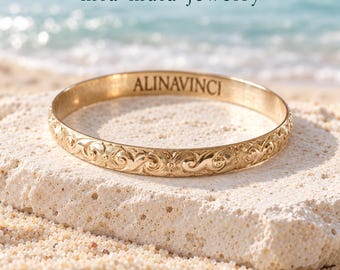 May include: A gold-toned metal bangle bracelet featuring an intricate engraved floral and scroll pattern along the exterior surface. The interior of the bracelet is engraved with the text ALINAVINCI. The brand name mia maia jewelry is displayed above the bracelet.