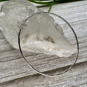 May include: A silver bangle bracelet with a textured design. The bracelet is on a white rock.