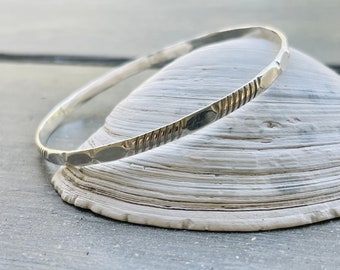 May include: A silver bangle bracelet with a textured design. The bracelet is on a white seashell.