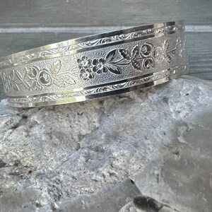 May include: A wide, silver-toned cuff bracelet with an engraved floral pattern. The bracelet is resting on a light gray rock, showcasing the intricate details of the design. The floral pattern includes flowers and leaves.