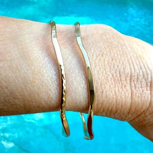 May include: Two gold-colored, hammered-style bracelets. The bracelets have a wavy, organic shape and are worn on a wrist. The background is a bright turquoise color.