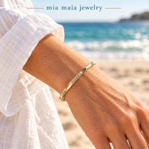 May include: A gold bangle bracelet featuring a repeating pattern of smooth, polished oval segments alternating with textured, twisted rope-style sections. The text mia maia jewelry is visible at the top of the image.