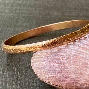 May include: A hammered gold bangle bracelet. The bracelet is made of a thick, solid piece of metal and has a smooth, polished finish.