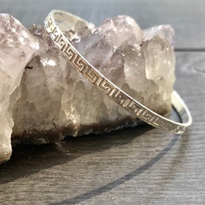 May include: A silver bangle bracelet with a Greek key design. The bracelet is on a white crystal surface.
