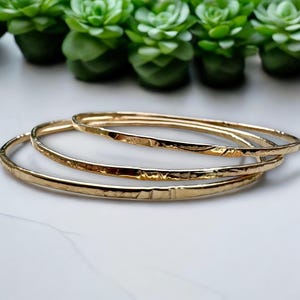 May include: Three gold-toned bangles stacked on top of each other. The bangles are all hammered and have a textured surface.