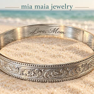 May include: A silver-toned metal bangle bracelet featuring an intricate embossed floral pattern along the outer edge and the engraved text "Love, Mom" on the inner surface. The brand name "mia maia jewelry" is visible above the bracelet.
