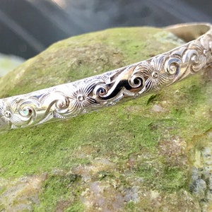 May include: A silver bangle bracelet with an intricate floral design. The bracelet is made of polished silver and has a delicate, feminine look.