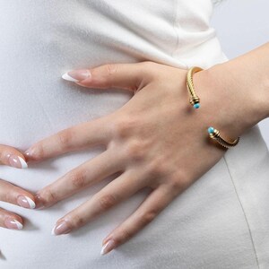 A person wearing a gold twisted cuff bracelet with turquoise ends on their wrist, viewed against a white top.