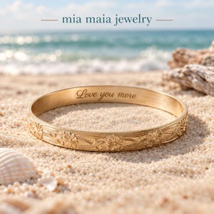 May include: A gold-toned metal bangle bracelet featuring an intricate embossed floral pattern along the exterior surface. The interior of the bracelet is engraved with the cursive text "Love you more." The brand name "mia maia jewelry" is visible.