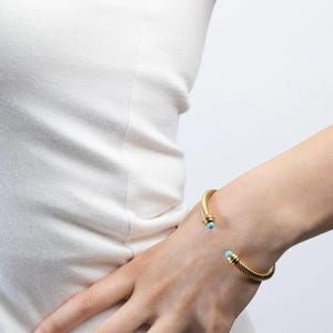 A person wearing a twisted gold cable cuff bracelet with small turquoise beads at both open ends on their wrist.