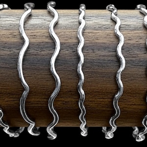 May include: Eight thin, silver-colored metal bangles, each featuring a continuous wavy or serpentine pattern. The bangles are arranged side-by-side, encircling a smooth, cylindrical wooden display rod with a visible dark brown wood grain texture.