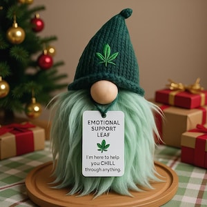 May include: A whimsical gnome with a green knitted hat and beard, holding a tag that reads "EMOTIONAL SUPPORT LEAF." The tag also includes the text "I'm here to help you CHILL through anything." The gnome is on a wooden tray, with a Christmas tree and wrapped gifts in the background.