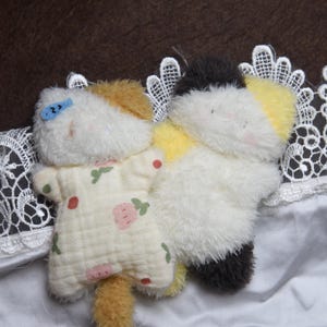 Peut inclure: Deux jouets en peluche en forme de chat. L'un des chats porte une tenue jaune clair avec un motif floral rose et rouge. L'autre chat est blanc, noir et jaune. Les deux jouets sont sur une surface blanche avec une bordure en dentelle.