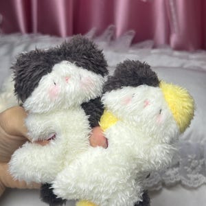 Peut inclure: Deux animaux en peluche avec des corps blancs, des fourrures noires et grises sur la t&ecirc;te et des joues roses. L'un a une queue jaune. Les jouets sont doux et moelleux et semblent faits &agrave; la main.