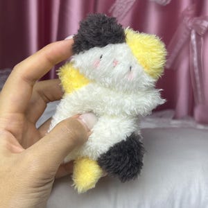 Peut inclure: Un petit chat en peluche blanc, jaune et noir. Le jouet a des joues roses et est tenu dans une main. L'arri&egrave;re-plan est un rideau rose tendre et un oreiller blanc.