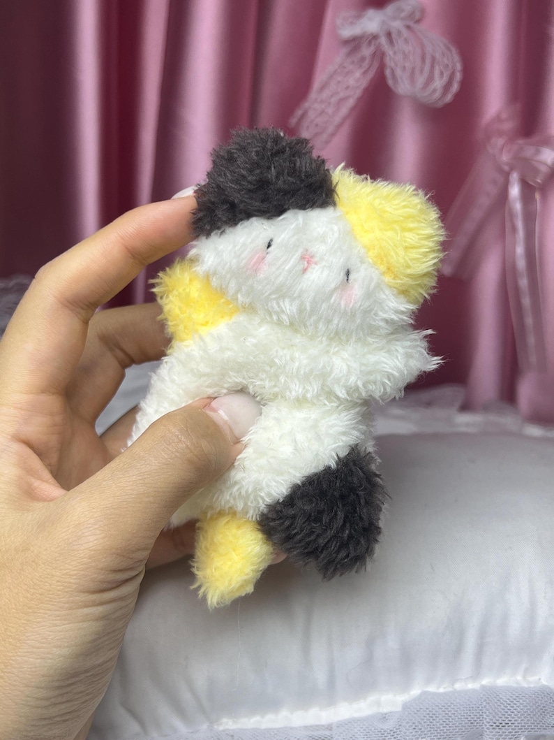 Peut inclure: Un petit chat en peluche blanc, jaune et noir. Le jouet a des joues roses et est tenu dans une main. L'arri&egrave;re-plan est un rideau rose tendre et un oreiller blanc.