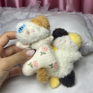 Peut inclure: Deux jouets en peluche en forme de chat. L'un est blanc avec des accents beiges et marron, portant une tenue &agrave; motifs floraux. L'autre est blanc avec des accents noirs et jaunes. Les deux ont des traits du visage brod&eacute;s et sont doux et duveteux.