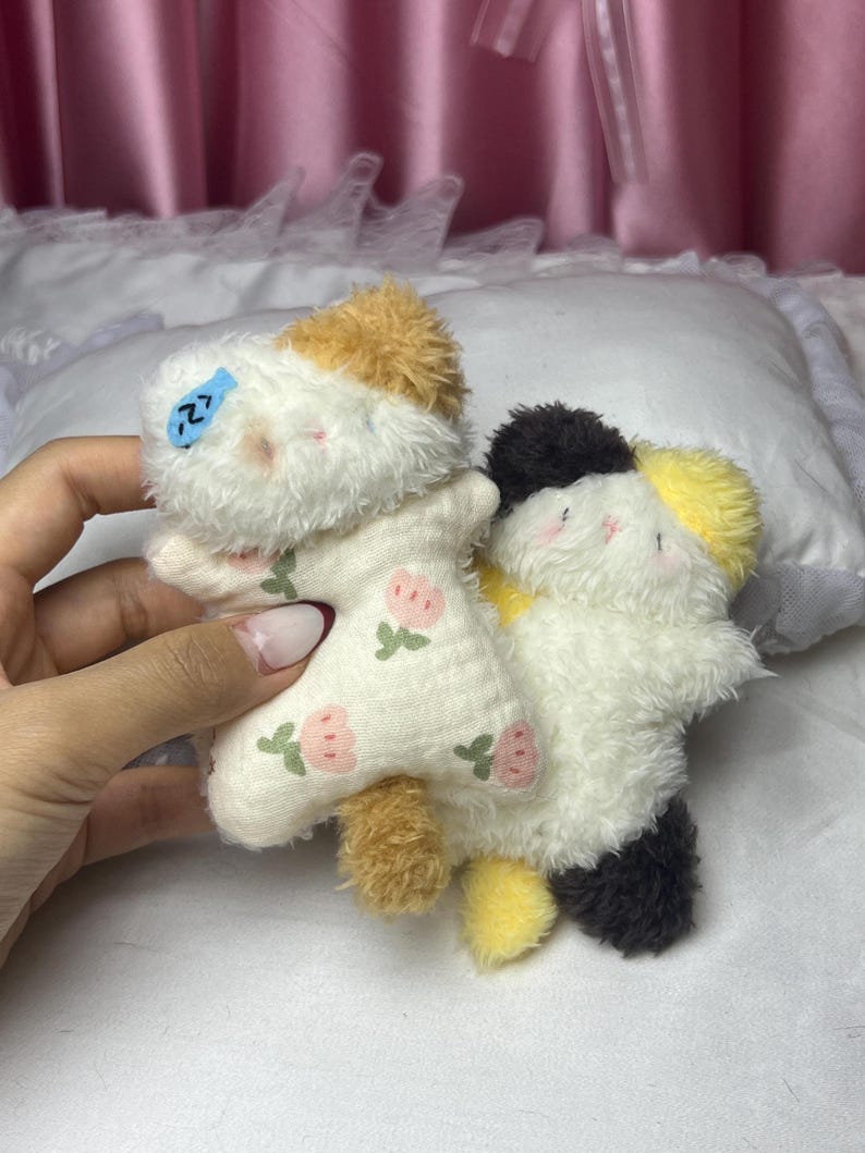Peut inclure: Deux jouets en peluche en forme de chat. L'un est blanc avec des accents beiges et marron, portant une tenue &agrave; motifs floraux. L'autre est blanc avec des accents noirs et jaunes. Les deux ont des traits du visage brod&eacute;s et sont doux et duveteux.