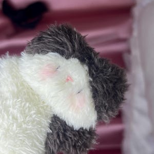 Peut inclure: Une peluche avec un design blanc et gris. Le jouet a une texture duveteuse, avec des accents roses sur les joues et les yeux ferm&eacute;s, sugg&eacute;rant un &eacute;tat de sommeil ou de d&eacute;tente. Le jouet est con&ccedil;u pour &ecirc;tre c&acirc;lin&eacute;.