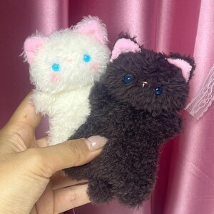 以下が含まれることがあります： 2つのぬいぐるみ猫のおもちゃ。片方はピンクの耳と青い目の白い猫、もう片方はピンクの耳と青い目の濃い茶色の猫です。どちらもふわふわした質感で、手で持たれています。
