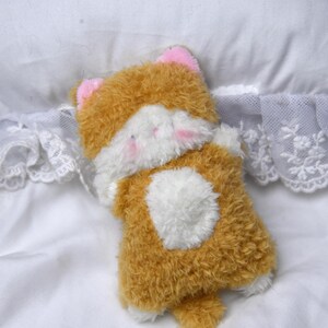 Peut inclure: Un jouet en peluche en forme de chat avec un corps marron et blanc, des oreilles roses et une poitrine blanche. Le jouet est pos&eacute; sur une surface blanche avec une bordure en dentelle.