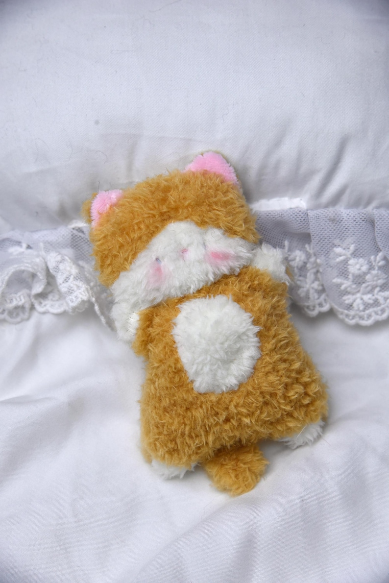 Peut inclure: Un jouet en peluche en forme de chat avec un corps marron et blanc, des oreilles roses et une poitrine blanche. Le jouet est pos&eacute; sur une surface blanche avec une bordure en dentelle.