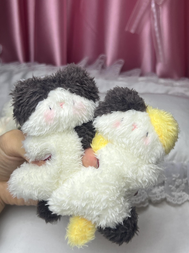 Peut inclure: Deux animaux en peluche avec des corps blancs, des fourrures noires et grises sur la t&ecirc;te et des joues roses. L'un a une queue jaune. Les jouets sont doux et moelleux et semblent faits &agrave; la main.