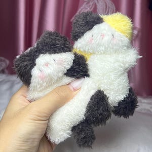 Peut inclure: Deux jouets en peluche en forme de chat. L'un est noir, blanc et jaune. L'autre est noir et blanc. Les jouets ont des joues roses et sont faits d'une mati&egrave;re douce et duveteuse. Les jouets sont tenus dans une main.