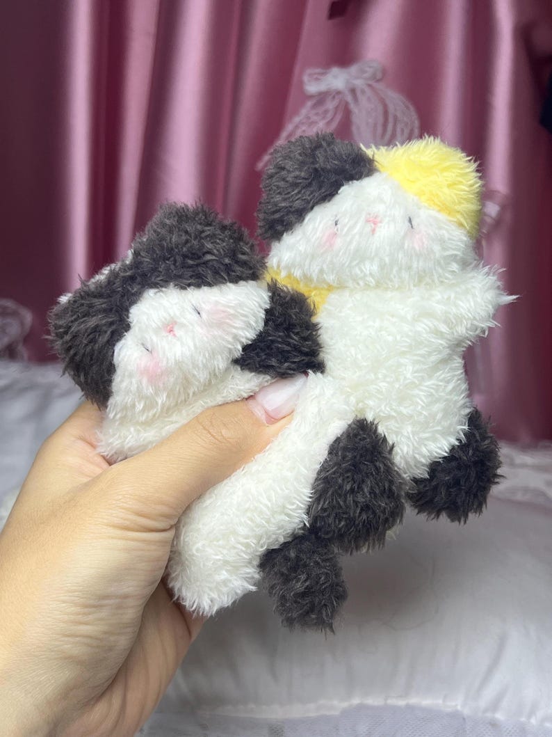 Peut inclure: Deux jouets en peluche en forme de chat. L'un est noir, blanc et jaune. L'autre est noir et blanc. Les jouets ont des joues roses et sont faits d'une mati&egrave;re douce et duveteuse. Les jouets sont tenus dans une main.