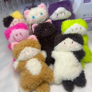 May include: A collection of handmade plush cat toys in various colors, including white, brown, black, purple, and green. Each plush cat has a fluffy texture, pink ears, and embroidered facial features. The toys are arranged together, showcasing their variety.