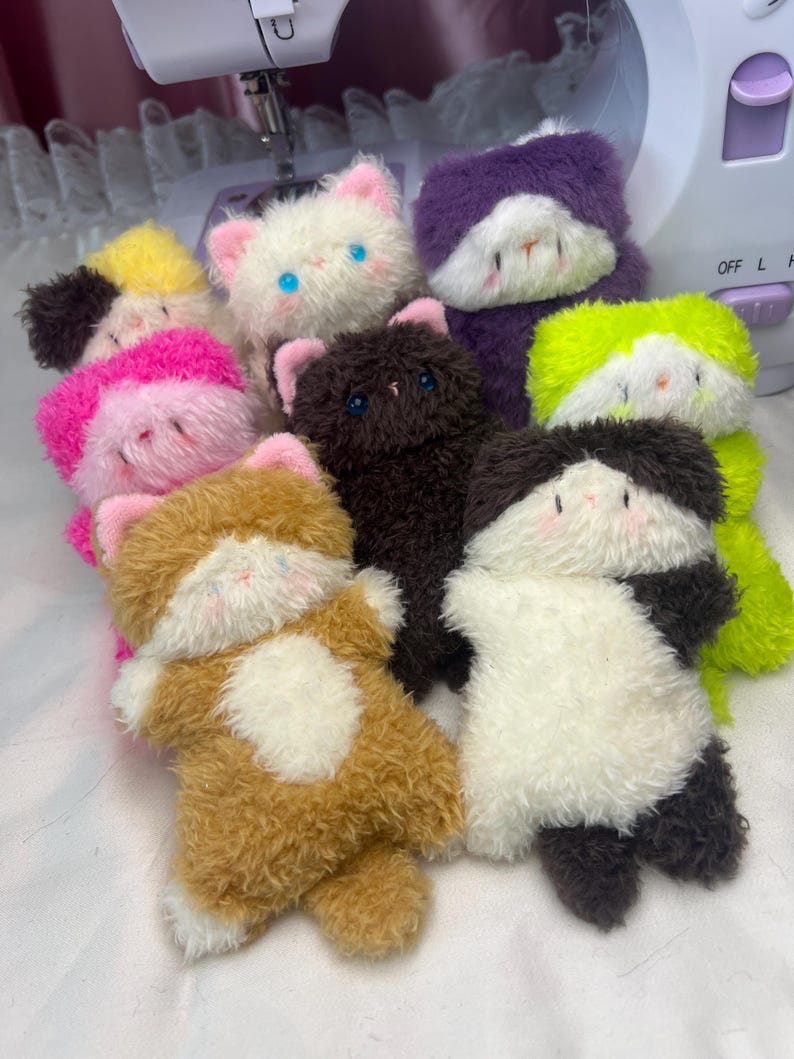 Peut inclure: Une collection de jouets en peluche faits &agrave; la main en forme de chat, de diff&eacute;rentes couleurs, dont blanc, marron, noir, violet et vert. Chaque chat en peluche a une texture duveteuse, des oreilles roses et des traits du visage brod&eacute;s.