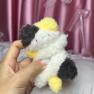 Peut inclure: Un jouet en peluche en forme de chat dans une pose c&acirc;line. Le jouet pr&eacute;sente un m&eacute;lange de fourrure blanche, jaune et noire. Le chat a de petites joues roses et est con&ccedil;u pour se donner l'impression de s'enlacer. Le jouet est doux et moelleux.