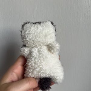 Peut inclure: Un petit jouet en peluche en forme d'animal duveteux. Le jouet est principalement blanc avec des accents gris fonc&eacute; sur la t&ecirc;te et une petite queue. Le jouet est tenu dans une main.