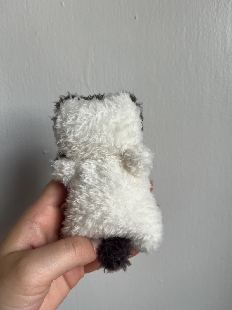 Peut inclure: Un petit jouet en peluche en forme d'animal duveteux. Le jouet est principalement blanc avec des accents gris fonc&eacute; sur la t&ecirc;te et une petite queue. Le jouet est tenu dans une main.