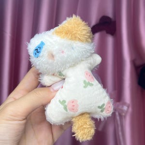 Peut inclure: Une petite peluche faite &agrave; la main en forme de chat. Le chat a un corps blanc avec une robe &agrave; motifs de fraises roses et de feuilles vertes. La t&ecirc;te du chat est blanche avec une touffe de fourrure beige et une petite queue beige. Un poisson bleu est brod&eacute; sur le visage du chat.