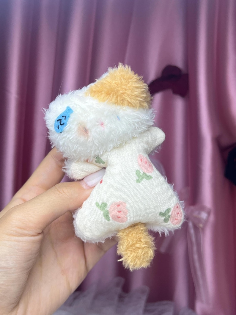 Peut inclure: Une petite peluche faite &agrave; la main en forme de chat. Le chat a un corps blanc avec une robe &agrave; motifs de fraises roses et de feuilles vertes. La t&ecirc;te du chat est blanche avec une touffe de fourrure beige et une petite queue beige. Un poisson bleu est brod&eacute; sur le visage du chat.