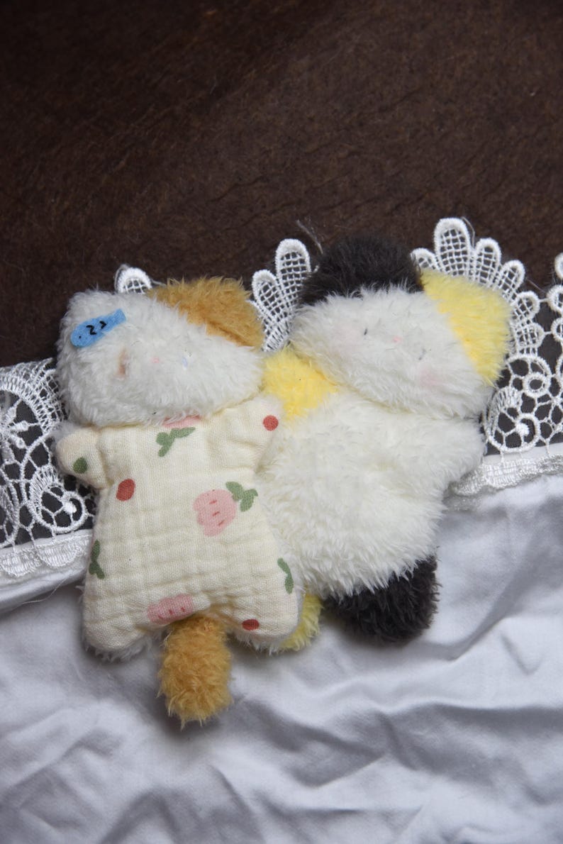 Peut inclure: Deux jouets en peluche en forme de chat. L'un des chats porte une tenue jaune clair avec un motif floral rose et rouge. L'autre chat est blanc, noir et jaune. Les deux jouets sont sur une surface blanche avec une bordure en dentelle.