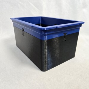 Puede incluir: Un contenedor de almacenamiento rectangular con borde azul y cuerpo negro. El contenedor tiene una superficie texturizada y peque&ntilde;os detalles decorativos en los laterales. El contenedor es de pl&aacute;stico duro.