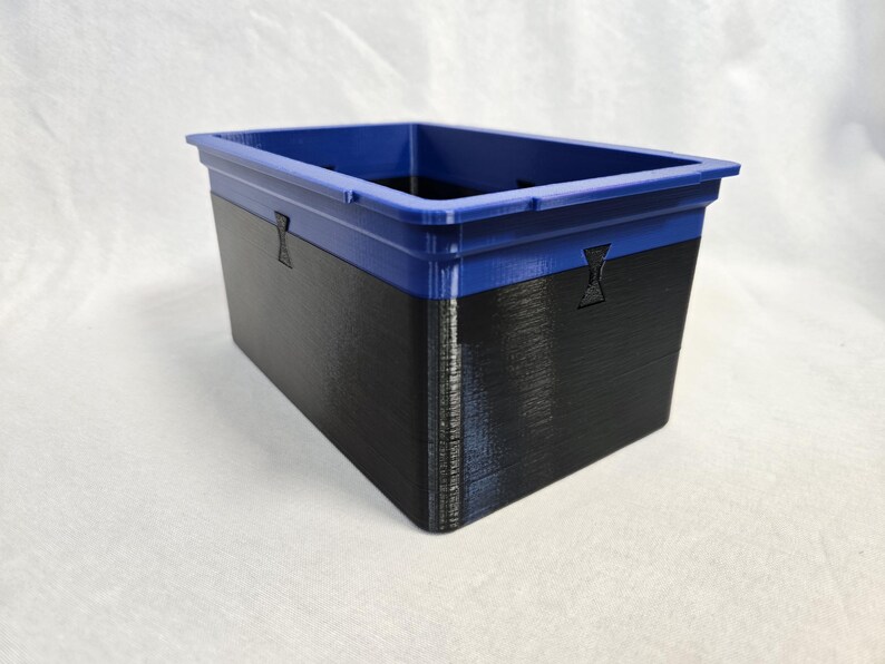 Puede incluir: Un contenedor de almacenamiento rectangular con borde azul y cuerpo negro. El contenedor tiene una superficie texturizada y peque&ntilde;os detalles decorativos en los laterales. El contenedor es de pl&aacute;stico duro.