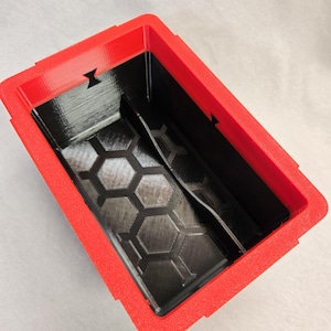 Puede incluir: Un contenedor rectangular rojo y negro con una superficie texturizada. El interior es negro con un patr&oacute;n de panal en la base. El marco rojo tiene detalles recortados en los lados.